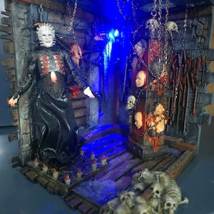 Può includere: Un diorama dettagliato che raffigura una figura di Pinhead in abito nero con spilli sulla testa, in una stanza buia con illuminazione blu. La scena include un pilastro decorato con volti umani, catene sospese e un mucchio di teschi.
