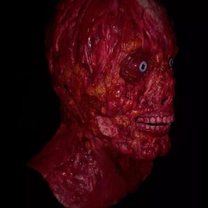 Pu&ograve; includere: Una maschera realistica e raccapricciante di una testa umana in decomposizione. La maschera &egrave; principalmente rossa, con carne e vene esposte e occhi blu. La bocca &egrave; aperta, rivelando i denti. Lo sfondo &egrave; nero.