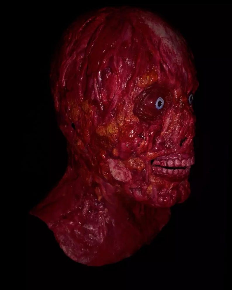 Pu&ograve; includere: Una maschera realistica e raccapricciante di una testa umana in decomposizione. La maschera &egrave; principalmente rossa, con carne e vene esposte e occhi blu. La bocca &egrave; aperta, rivelando i denti. Lo sfondo &egrave; nero.