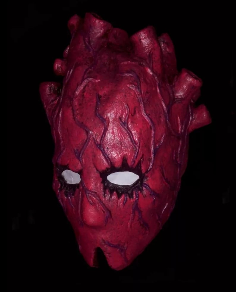 Peut inclure: Un masque rouge en forme de c&oelig;ur humain, avec des ouvertures blanches pour les yeux et des veines violet fonc&eacute;. Le masque a une surface textur&eacute;e et est pos&eacute; sur un fond noir. Accessoire de costume unique.