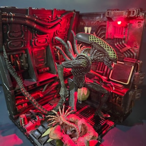 Puede incluir: Diorama detallado con una figura de Alien Xenomorfo con un cuerpo texturizado y una cabeza distintiva. La escena incluye un 'chestburster' emergiendo de un huevo, todo en un entorno industrial oscuro con iluminación roja.