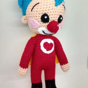Pu&ograve; includere: Bambola all'uncinetto fatta a mano in costume da supereroe. La bambola ha un corpo rosso, stivali neri e un mantello giallo. Il viso &egrave; beige con capelli blu, naso rosso e sorriso bianco. Un cerchio bianco con un cuore rosso &egrave; sul petto.