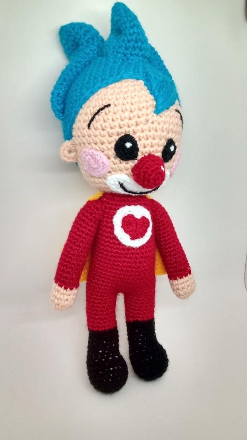 Pu&ograve; includere: Bambola all'uncinetto fatta a mano in costume da supereroe. La bambola ha un corpo rosso, stivali neri e un mantello giallo. Il viso &egrave; beige con capelli blu, naso rosso e sorriso bianco. Un cerchio bianco con un cuore rosso &egrave; sul petto.