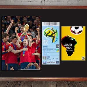 May include: Framed display featuring a photo of a soccer team celebrating a victory, a ticket, and a yellow poster with the text "2010 FIFA WORLD CUP SOUTH AFRICA". The photo shows players in red jerseys holding a trophy.