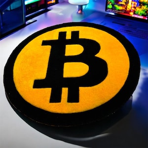 May include: Round rug featuring the Bitcoin symbol. The rug has a yellow center with a large black "B" and a black border. The rug is circular and appears to be made of a soft material.
