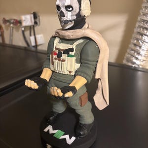 May include: A figurine of a character from the video game Call of Duty: Modern Warfare. The figure has a skull mask, headset, and military-style clothing. The base is black with the letters "MW" in white and green.