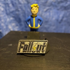 May include: A silver-colored Fallout pin with the game's logo and a yellow lightning bolt. A small figurine of the Vault Boy character, wearing a blue jumpsuit with yellow accents, stands behind the pin.