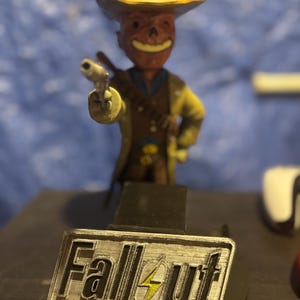 May include: A figurine of a cowboy-themed character from the Fallout video game series. The figure is holding a gun and wearing a yellow hat and coat. The base features the Fallout logo.