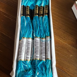 Puede incluir: Una caja de seis carretes de hilo de bordar azul turquesa y blanco. Los carretes están etiquetados con "Iris Rayon 100%" y tienen un código de barras y el número "600" impreso en ellos.