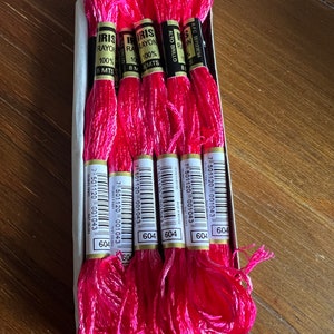 Op de afbeelding: Een doos met 8 spoelen roze borduurgaren, elke spoel met het label "Iris Rayon 100% Alto Brillo 8 MTS", een barcode en het nummer "604".