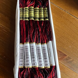 May include: Six skeins of Iris Rayon embroidery thread in a variegated red and dark burgundy colorway, packaged in a white cardboard box. Each skein features a label with the text Iris Rayon 100% 8 MTS, a barcode, and the number 608.