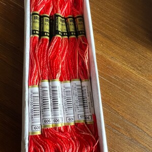 Op de afbeelding: Een doos met zes spoelen rode en witte borduurgaren. De spoelen zijn gelabeld met "IRIRA RAY" en "ALTO BRILLO" met het nummer "606" erop gedrukt.