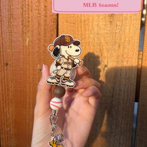 May include: A retractable badge reel featuring a Snoopy character wearing a brown and white San Diego Padres baseball uniform and cap, holding a bat. Below are brown and white beads, a baseball-patterned bead, and a yellow Woodstock charm.