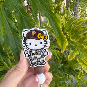 May include: A badge reel featuring a Hello Kitty character wearing a brown and gold San Diego Padres baseball cap and a pinstriped jersey, holding a baseball. The reel includes a brown bead and a white baseball-themed bead above the clip.