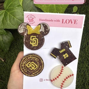 May include: Four decorative pins on a white display card. One is a brown glittery mouse head with a yellow bow and SD logo, one is a round brown glittery San Diego Padres logo, one is a brown jersey with Tatis 23, and one is a white baseball.