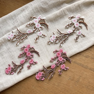 May include: Four embroidered patches featuring cherry blossom branches. Two patches display white and light pink flowers on brown stems, while the other two feature vibrant pink flowers on brown stems, all with detailed stitching and thread work.