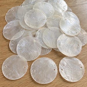 Peut inclure: Un ensemble de disques circulaires en nacre blanche translucide, chacun doté de deux petits trous pré-percés sur les côtés opposés. Ces composants plats et fins présentent une surface naturelle, irisée et légèrement texturée.