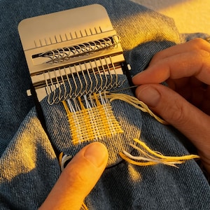 May include: A close-up of a metal weaving tool in use. The tool is gold-colored and has multiple hooks and a sliding mechanism. Yellow and white thread is being woven into a denim fabric, creating a small patch. The hands of a person are visible.