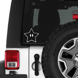 May include: White vinyl decal of a cartoon star with two small black oval eyes. The star is affixed to the rear window of a white vehicle, near the tyre and taillight.