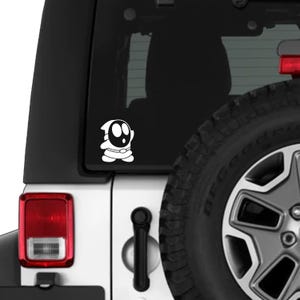 Könnte beinhalten: Weißer Vinyl-Aufkleber eines Shy Guy-Charakters aus der Super Mario-Videospielserie. Der Aufkleber ist auf der Heckscheibe eines weißen Fahrzeugs angebracht. Die Figur hat eine schwarze Maske und einen weißen Körper.