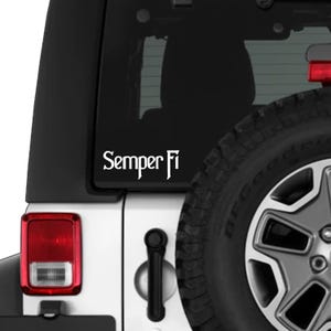 Pode incluir: Autocolante de vinil branco com as palavras "Semper Fi" numa fonte estilizada, afixado no vidro traseiro de um veículo branco. A imagem inclui uma porção de um pneu preto e uma luz traseira vermelha.