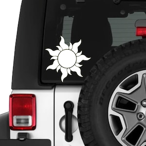 May include: White vinyl decal of a stylized sun with pointed rays, affixed to the rear window of a white vehicle. The sun design is centered, with a black background visible through the window. A portion of a tire is visible.