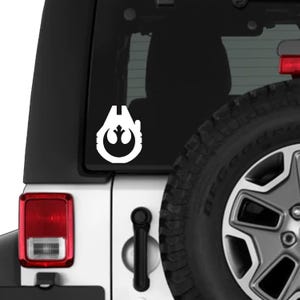 Puede incluir: Calcomanía de vinilo blanco con el símbolo de la Alianza Rebelde, adherida a la parte trasera de un vehículo blanco. El emblema es un diseño estilizado, un círculo con un motivo central, y está colocado cerca de la ventana trasera y el neumático.