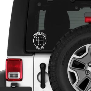 May include: White vinyl decal featuring a gear shift pattern with the words "ENDANGERED SPECIES". The decal is circular and is affixed to the rear window of a white vehicle. The vehicle's tire and tail light are also visible.