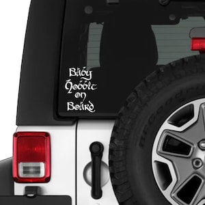 Op de afbeelding: Witte vinyl sticker met de tekst "Baby Hobbit on Board" in een grillige lettertype. De sticker is bevestigd op de achterruit van een witte Jeep. De afbeelding bevat een deel van de band en het achterlicht.