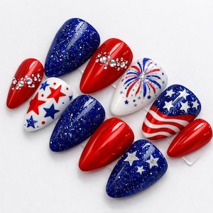 May include: A collection of artificial nails in red, white, and blue, featuring various designs. Some nails are solid colors, while others have glitter, star patterns, or rhinestone accents. One nail showcases a firework design.