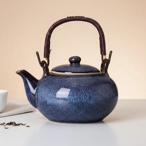 Könnte beinhalten: Eine blaue Keramik-Teekanne mit einem geflochtenen braunen Griff und einem passenden Deckel. Die Teekanne hat einen Ausguss und einen kleinen Knopf auf dem Deckel. Der Hintergrund ist in einem weichen Beigeton gehalten, und die Teekanne steht auf einer weißen Oberfläche.