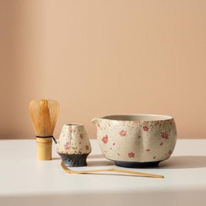 May include: A matcha tea set featuring a bamboo whisk, a ceramic bowl with a spout, a small ceramic holder, and a bamboo scoop. The bowl and holder have a speckled beige finish with red floral accents. The whisk has a natural wood color.