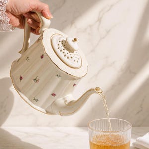 Puede incluir: Tetera de porcelana color crema con diseño floral y ribetes dorados, vertiendo líquido en un vaso transparente. La tetera tiene una tapa decorativa con pequeños agujeros y un asa curvada. El líquido parece ser té.