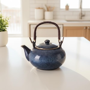Może przedstawiać: Niebieski ceramiczny czajnik z plecionym brązowym uchwytem i pasującą pokrywką. Czajnik ma zaokrąglony kształt i dzióbek z boku. Czajnik stoi na białej powierzchni.