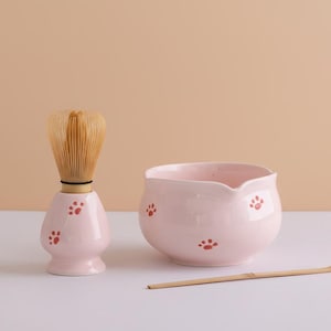 Puede incluir: Un juego de cuenco y batidor de matcha de cerámica rosa. El cuenco y el soporte del batidor están decorados con huellas de patas rojas. El batidor es de bambú. El juego está diseñado para preparar té matcha.