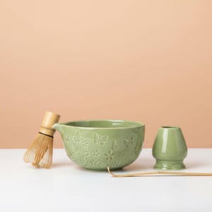 May include: A matcha tea set featuring a light green ceramic bowl with butterfly and floral designs, a bamboo whisk, a small green ceramic tea cup, and a bamboo tea scoop. The set is on a white surface with a peach background.