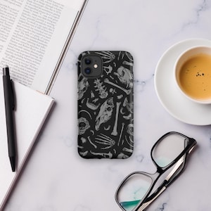 May include: Black phone case featuring a repeating pattern of gray bones, skulls, and skeletal elements. The case is shown on a marble surface, next to a cup of coffee and a pair of glasses.