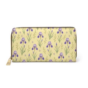 May include: A rectangular zip-around wallet featuring a pale yellow background with a repeating pattern of purple iris flowers, green stems, and small fleur-de-lis symbols. The wallet has a gold-toned metal zipper pull attached to the side.