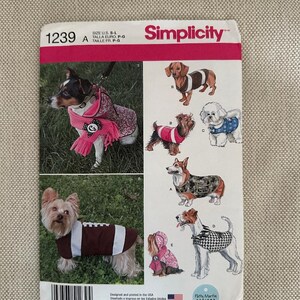 May include: Simplicity pattern 1239 A for dog clothing. The pattern features various dog breeds wearing different outfits, including a football-themed coat and a pink hooded jacket. The pattern size is S-L.