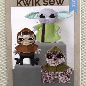 May include: A Kwik Sew pattern for 18-inch plush aliens. The pattern features three different alien designs, including a green-robed character with large ears, a brown-toned alien with a hood, and another with a brown cowl.