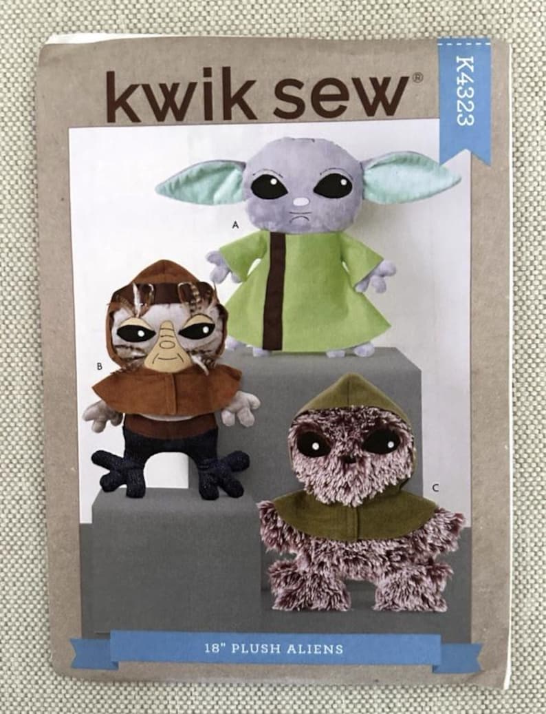 May include: A Kwik Sew pattern for 18-inch plush aliens. The pattern features three different alien designs, including a green-robed character with large ears, a brown-toned alien with a hood, and another with a brown cowl.