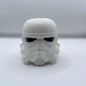 May include: A white 3D printed figurine of a stormtrooper helmet wrapped in bandages. The helmet features a smooth matte finish with distinct layered lines from the printing process, dark eye lenses, and a textured mummy-like aesthetic.