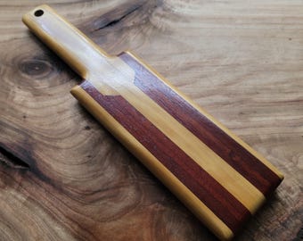 Op de afbeelding: Een rechthoekige houten peddel met een handvat dat aan het uiteinde een gat heeft. Het oppervlak is opgebouwd uit afwisselende verticale stroken lichtbruin en donker roodbruin hout, afgewerkt met een gladde, glanzende coating.