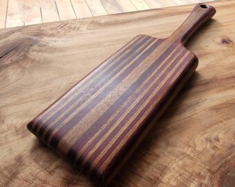 Op de afbeelding: Een rechthoekige houten serveerplank met een gestreept patroon van afwisselend donkerpaarse en lichtbruine houtnerven. De plank heeft een lang, geïntegreerd handvat met een klein rond gat aan het uiteinde voor ophangopslag.