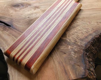Op de afbeelding: Een rechthoekige houten serveerplank met afwisselende verticale strepen van lichtbruin en donker roodbruin hout. De plank heeft een lang, geïntegreerd handvat met een klein rond gat aan het uiteinde om de plank aan op te hangen.