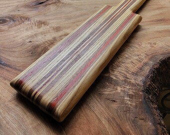Op de afbeelding: Een rechthoekige houten serveerplank met een gestreept patroon van lichtbruine, donkerbruine en roodachtige houtlagen. De plank heeft een lang, smal handvat met een klein rond gat aan het uiteinde om de plank aan op te hangen.