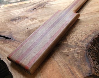 Op de afbeelding: Een rechthoekig houten serveerplank met afwisselende verticale strepen in lichtbruin, donkerbruin en roodachtige tinten. Het handvat heeft een klein rond gat aan het uiteinde om op te hangen en het oppervlak heeft een gladde afwerking.