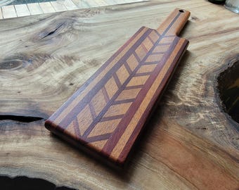 Op de afbeelding: Een rechthoekige houten serveerplank met een visgraatpatroon van afwisselend lichte en donkere houten stroken. De plank heeft een handvat met een klein rond gat aan de bovenkant en een gladde, gepolijste afwerking met houtnerf.