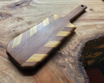 Op de afbeelding: Een rechthoekige houten serveerplank met een handvat met een gat aan het uiteinde. De plank is gemaakt van afwisselende diagonale stroken donkerbruin walnotenhout en lichter hout, wat een visgraatpatroon op het oppervlak creëert.