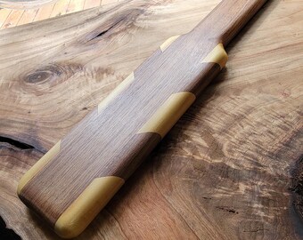 Op de afbeelding: Een lange, platte houten peddel met een donkerbruine houtnerf en contrasterende lichtbruine houten accenten op de hoeken en randen. Het handvat heeft een klein rond gat aan het uiteinde om het voorwerp op te hangen.